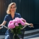 Lächelnde Frau auf einem Fahrrad mit einem Korb voller blühender rosa Pfingstrosen vor einer dunklen, wellenförmigen Wand an einem sonnigen Tag.